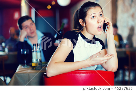 Offended young woman speaking by phone on background with drunk man at restaurant table 45287635