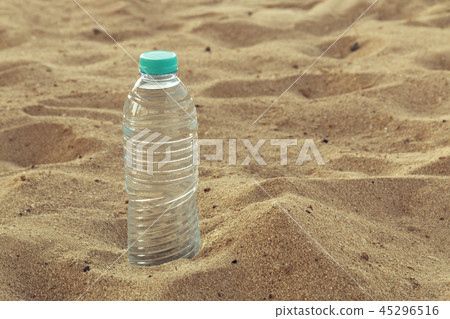 Bottle with crystal clear drinking water with a red lid on the sand on a hot, sunny day closeup. Bottle with crystal clear drinking water with a red lid on the sand on a hot, sunny day closeup. 45296516