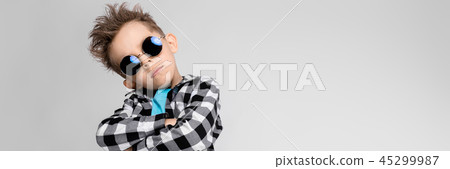 A handsome boy in a plaid shirt, blue shirt and jeans stands on a gray background. The boy is 45299987