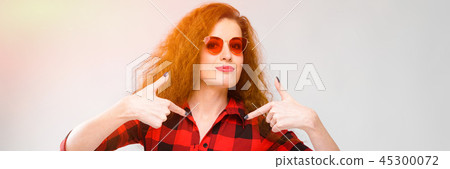 Portrait of beautiful redhead happy young woman in sunglasses smiling showing on blank area on gray Portrait of beautiful redhead happy young woman in sunglasses smiling showing on blank area on gray 45300072