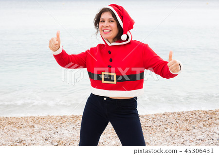 Christmas, humor and people concept - young happy woman in Santa Claus suit at the beach showing Christmas, humor and people concept - young happy woman in Santa Claus suit at the beach showing 45306431