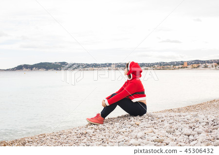 Sea, holidays and christmas concept - young funny woman in santa costume sitting on a beach 45306432