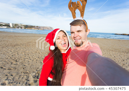 Fun and winter holidays concept - Happy couple in christmas costumes taking selfie over sandy beach Fun and winter holidays concept - Happy couple in christmas costumes taking selfie over sandy beach 45307475