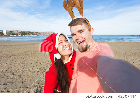 Christmas, humor, people concept - Happy Christmas couple having fun taking selfie on beach Christmas, humor, people concept - Happy Christmas couple having fun taking selfie on beach 45307484