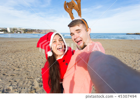 Fun and winter holidays concept - Happy couple in christmas costumes taking selfie over sandy beach Fun and winter holidays concept - Happy couple in christmas costumes taking selfie over sandy beach 45307494