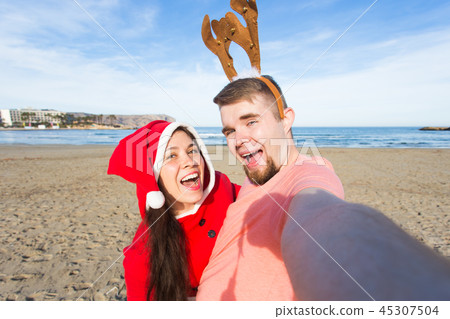 Christmas, humor, people concept - Happy Christmas couple having fun taking selfie on beach Christmas, humor, people concept - Happy Christmas couple having fun taking selfie on beach 45307504
