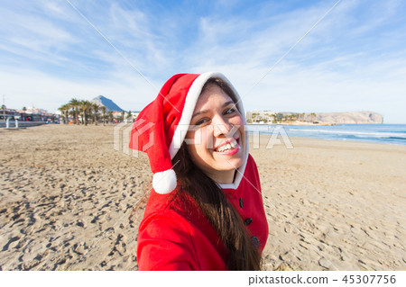 People, holidays and christmas concept - young woman in santa costume taking selfie on beach 45307756