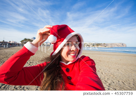 People, holidays and christmas concept - young woman in santa costume taking selfie on beach People, holidays and christmas concept - young woman in santa costume taking selfie on beach 45307759