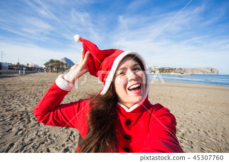 People, holidays and christmas concept - young woman in santa costume taking selfie on beach People, holidays and christmas concept - young woman in santa costume taking selfie on beach 45307760