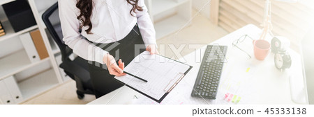 A beautiful young girl stands near an office desk and holds in her hands a sheet for notes and a 45333138