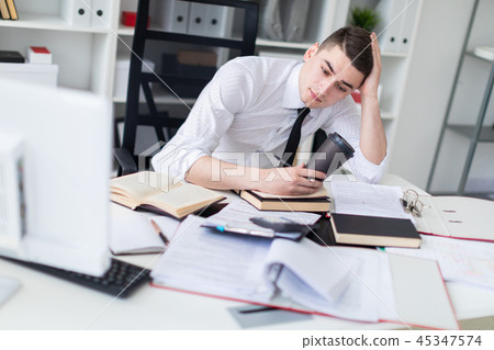 A young man working at a table in the office with a book, documents and a computer. He is holding a 45347574