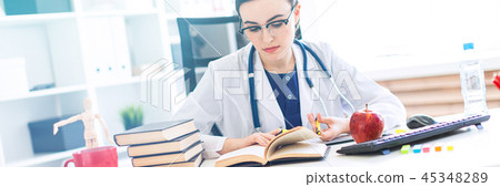 A beautiful young girl in a white robe sits at a computer desk, holds a marker and a pen in her hand 45348289