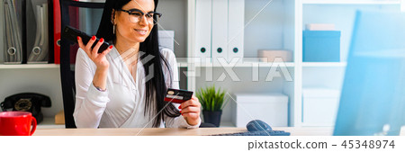 A young girl is sitting at the table, holding a bank card and phone. A young girl is sitting at the table, holding a bank card and phone. 45348974
