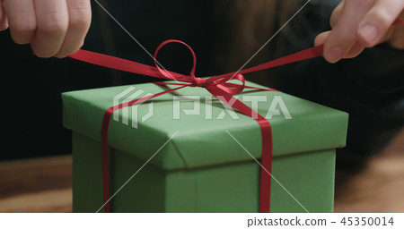 closeup shot of young female hands unties red ribbon bow on green paper gift box 45350014