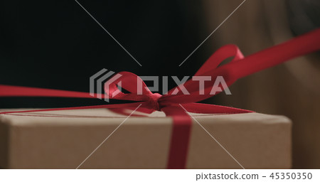 closeup shot of young female hands unties red ribbon bow on craft paper gift box closeup shot of young female hands unties red ribbon bow on craft paper gift box 45350350