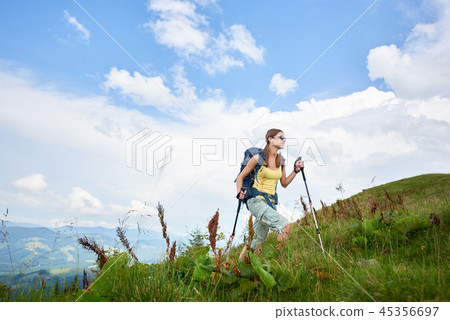 Woman hiker hiking on grassy hill, wearing backpack, using trekking sticks in the mountains 45356697