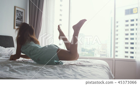 Happy woman jumping on bed at home and relaxing by the panoramic window with city view 45373066