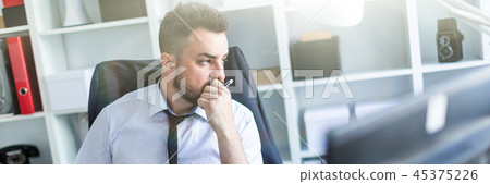 A man sits in a chair in the office and holds a pencil in his hand. On the back of the chair hangs 45375226