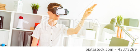 A young man in virtual reality glasses stands near the table and pulls his arm up. A young man 45375269