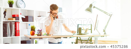 A young man in glasses stands near a computer desk and is talking on the phone. Before him lies a 45375286