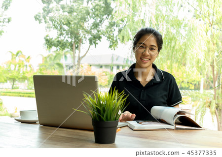Asian woman with glasses smiling and open a book with laptop on table in cafe. Asian woman with glasses smiling and open a book with laptop on table in cafe. 45377335