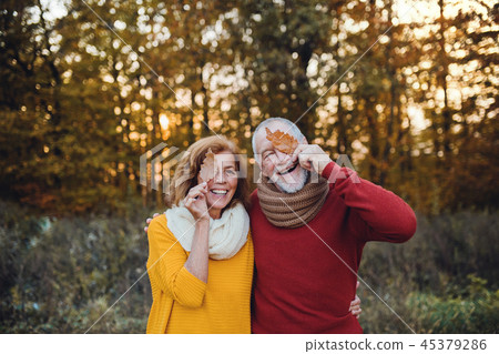 A senior couple standing in an autumn nature at sunset, covering eyes with leaves. A senior couple standing in an autumn nature at sunset, covering eyes with leaves. 45379286