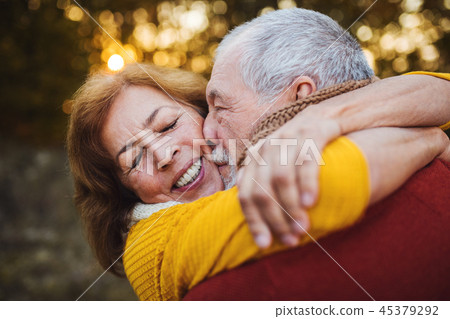A senior couple standing in an autumn nature at sunset, hugging and kissing. 45379292
