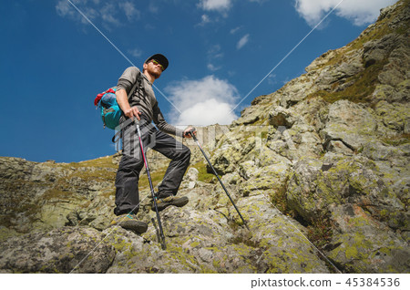 A bearded man in sunglasses and a cap with a backpack stands on top of a rock and looks into a rocky A bearded man in sunglasses and a cap with a backpack stands on top of a rock and looks into a rocky 45384536