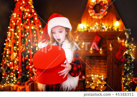 Adorable little girl opening a magical Christmas gift by a Christmas tree in cozy living room in Adorable little girl opening a magical Christmas gift by a Christmas tree in cozy living room in 45388351