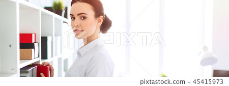 A young girl in the office is standing near the shelf and pulling out a red folder. 45415973