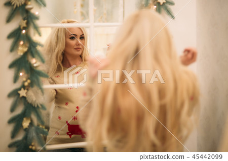A pretty pregnant blonde hair woman in a Christmas sweater stands near the mirror. Winter spirit 45442099
