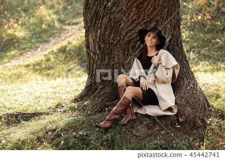 woman in a beige coat and black hat sitting under tree woman in a beige coat and black hat sitting under tree 45442741