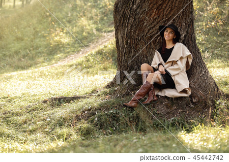 woman in a beige coat and black hat sitting under tree woman in a beige coat and black hat sitting under tree 45442742