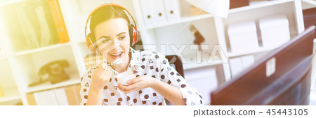 Beautiful young girl sitting in headphones at desk in office, eating yogurt and looking at monitor. 45443105