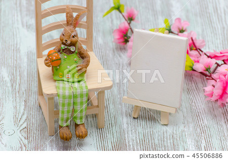 Easter bunny sitting on the stool with a egg, painting easel and flowers over wooden background 45506886