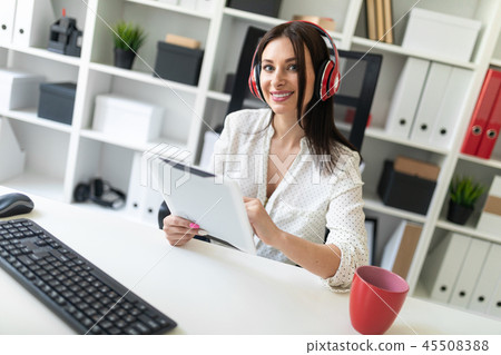 A young girl with headphones sitting in the office at the computer table and holding a tablet. A young girl with headphones sitting in the office at the computer table and holding a tablet. 45508388