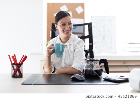 A young girl sitting at a table in the office and holding a Cup. A young girl sitting at a table in the office and holding a Cup. 45521869