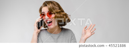 Charming young girl in a gray T-shirt on a gray background. Girl in red glasses with phone 45521886