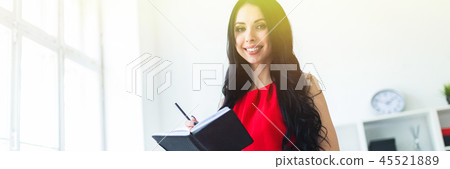 Beautiful young girl in a red suit is standing in the office and holding a notebook and a pencil. Beautiful young girl in a red suit is standing in the office and holding a notebook and a pencil. 45521889