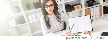 A beautiful young girl is sitting at a table in the office and pointing a pencil at the information A beautiful young girl is sitting at a table in the office and pointing a pencil at the information 45521895