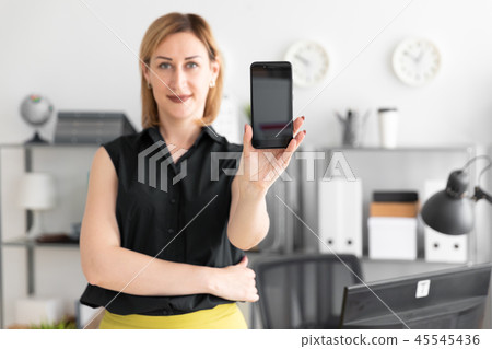 Young girl standing in the office and shows the phone screen. Young girl standing in the office and shows the phone screen. 45545436