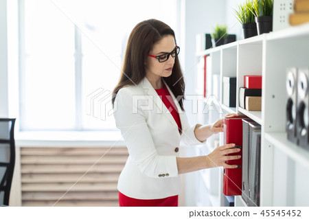 A young girl stands in the office near the rack and puts a folder on it. 45545452