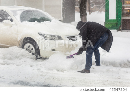 Car blocked with snow drift on city street. Man cleaning vehicle from snow with brush during heavy Car blocked with snow drift on city street. Man cleaning vehicle from snow with brush during heavy 45547204