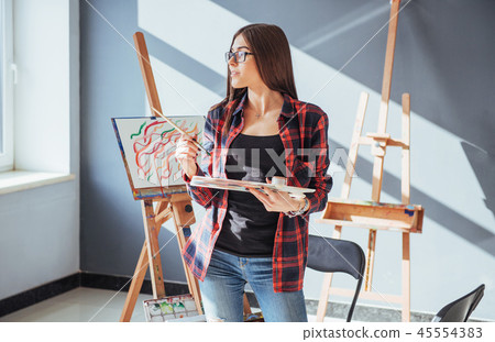 Creative pensive painter girl paints a colorful picture on canvas with oil colors in workshop. 45554383