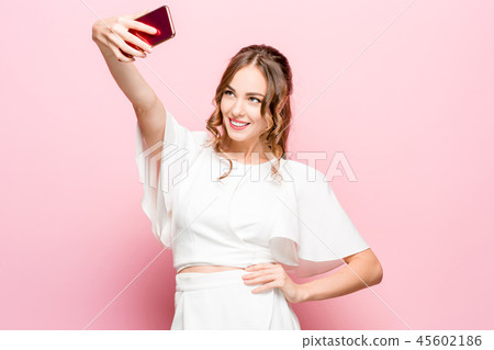 Portrait of a young attractive woman making selfie photo with smartphone on a pink background 45602186