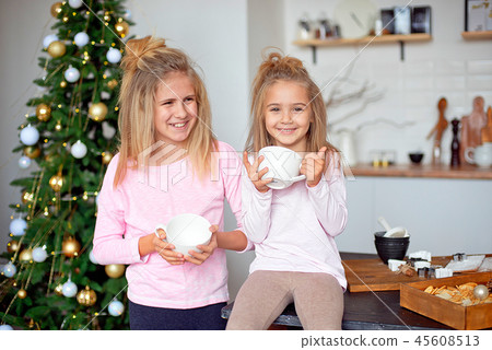 Two sisters in their pajamas in the kitchen drinking tea from big white cup on the background of the 45608513