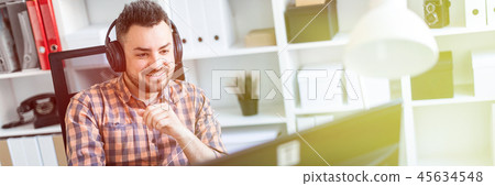 A young man in headphones sits at a table in the office and looks at the monitor. A young man in headphones sits at a table in the office and looks at the monitor. 45634548