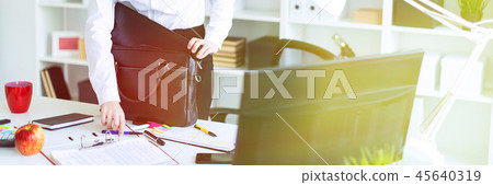 A young girl in the office is standing near the table and puts things in a bag. 45640319