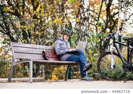 A senior man with electrobike sitting on a bench outdoors in town, using laptop. 45663718