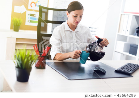 A young girl sitting at a table in the office and pours coffee into a Cup. A young girl sitting at a table in the office and pours coffee into a Cup. 45664299